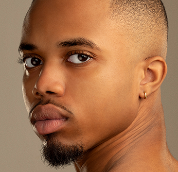 A close-up photo of a person with a short shaved haircut, a small hoop earring in the left ear, and a neatly trimmed goatee. The individual has a serious expression and is looking directly at the camera with a neutral, beige background.