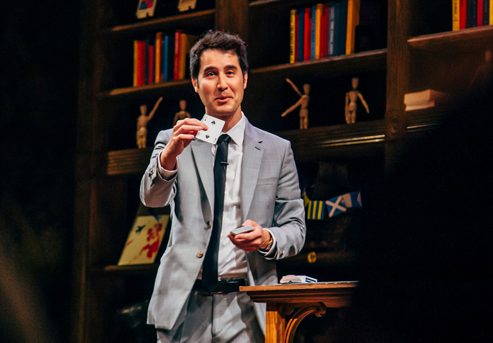 A man in a gray suit holds up a playing card in front of bookshelves filled with books, figurines, and decorations.