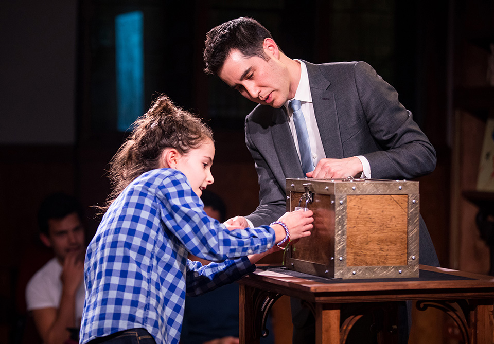 A young girl in a checkered shirt works on a lock on a wooden box as a man in a suit observes.