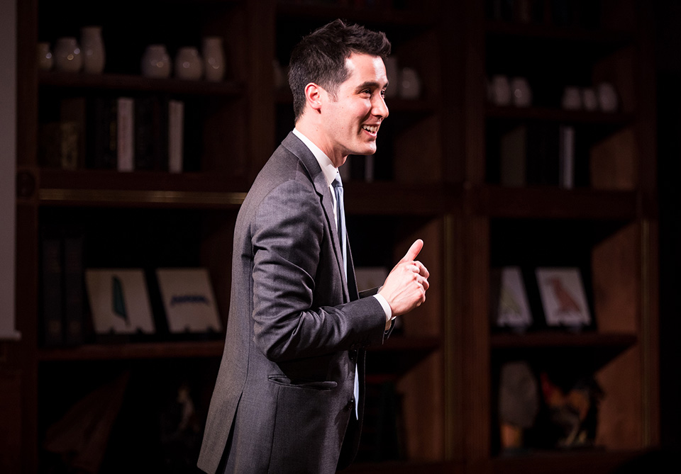 Man in a gray suit speaking in front of wooden bookshelves with books and framed pictures.