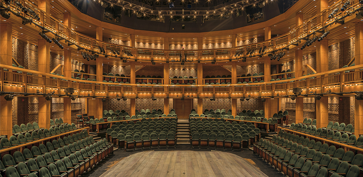 Theater interior with three levels of seating, green upholstered chairs, and wooden architectural details.