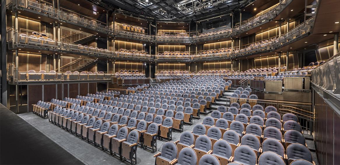 Interior view of a large, modern multi-tiered theater from the stage, with blue cushioned chairs and warm lighting.