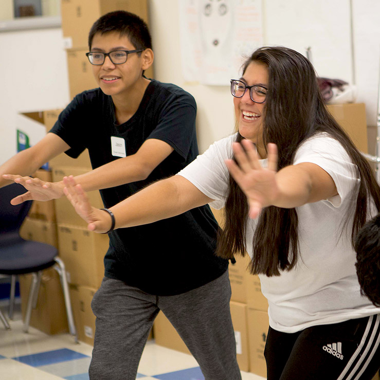 Two kids smiling and extending their hands forward in a classroom.