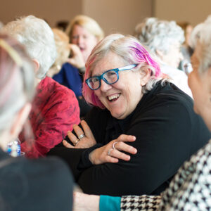 Woman with pink-streaked gray hair and blue glasses laughing in a social gathering.