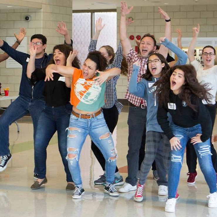 A group of eleven people indoors, energetically posing with raised arms and open mouths.