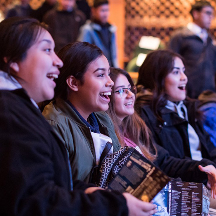 Four young women sitting close together in a theater, smiling, holding programs, and looking at the stage off-camera. 