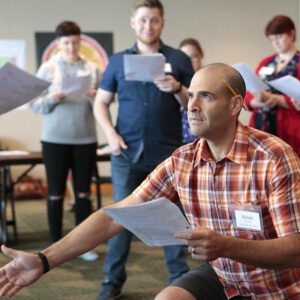 Man wearing a plaid shirt, identified as Kevin, kneeling and holding a paper, surrounded by four people standing and holding papers.