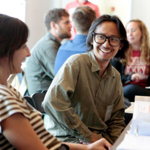 A man with black hair in glasses smiling and talking to a woman in a striped shirt, with others talking in the background.