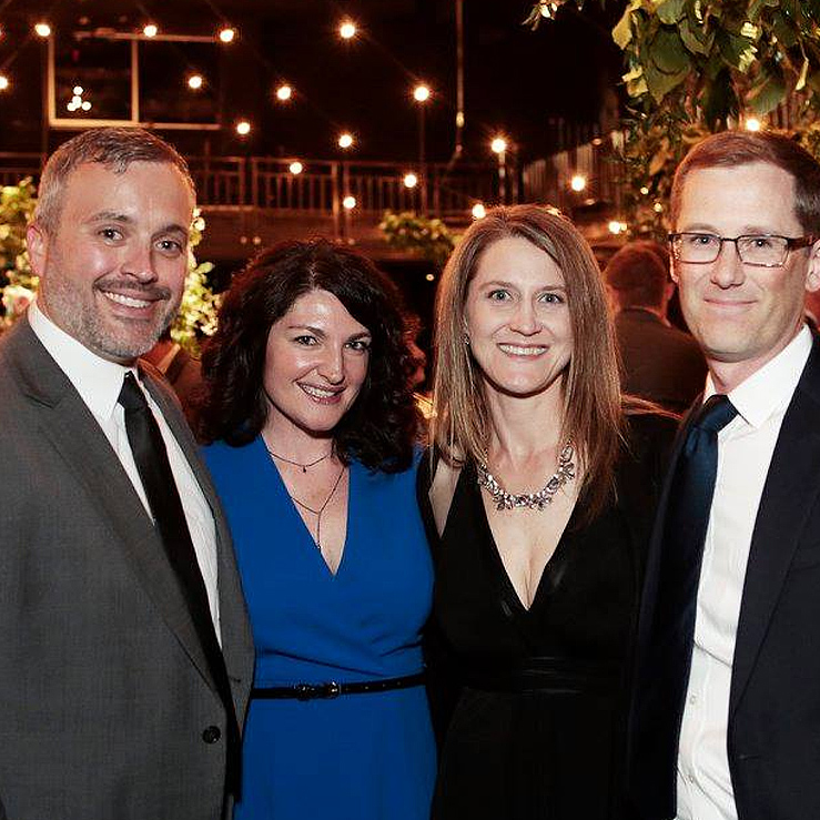 Four people smiling at a festive indoor event with string lights and greenery in the background.