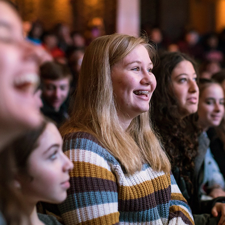 A group of young people seated and smiling in a theater, with a girl in a striped sweater and braces in the center.
