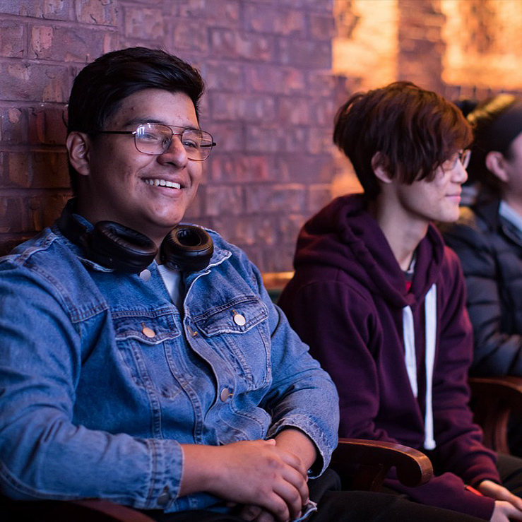 Three young people sitting against the brick wall of the back of the theater, one of them smiling, wearing a denim jacket and headphones, another in a maroon hoodie, and the last in a black jacket.