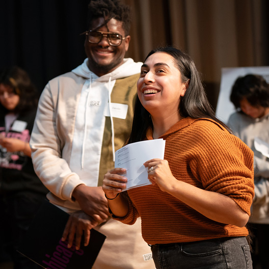 Two people smiling at an event, one holding a piece of white paper, others in the background.