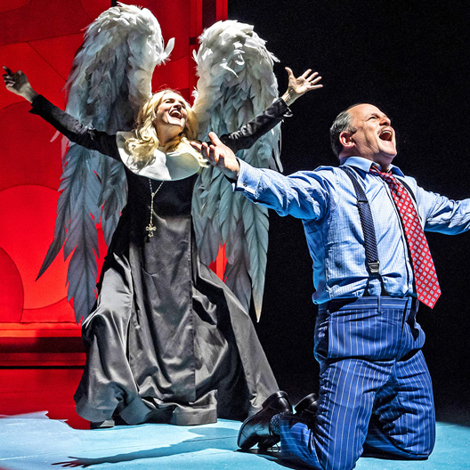 Two performers in a dramatic theatrical scene, one dressed as an angel with white wings and a black robe, and the other in a blue pinstripe suit kneeling, both with expressive and animated poses.