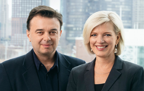 Two people in professional attire, Artistic Director Edward Hall (on left) and Executive Director Kim Motes (on right), standing in front of a blurred cityscape.