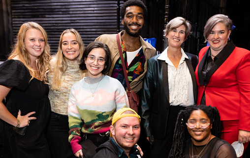  A group of eight people posing and smiling at the camera indoors with a wooden wall background.