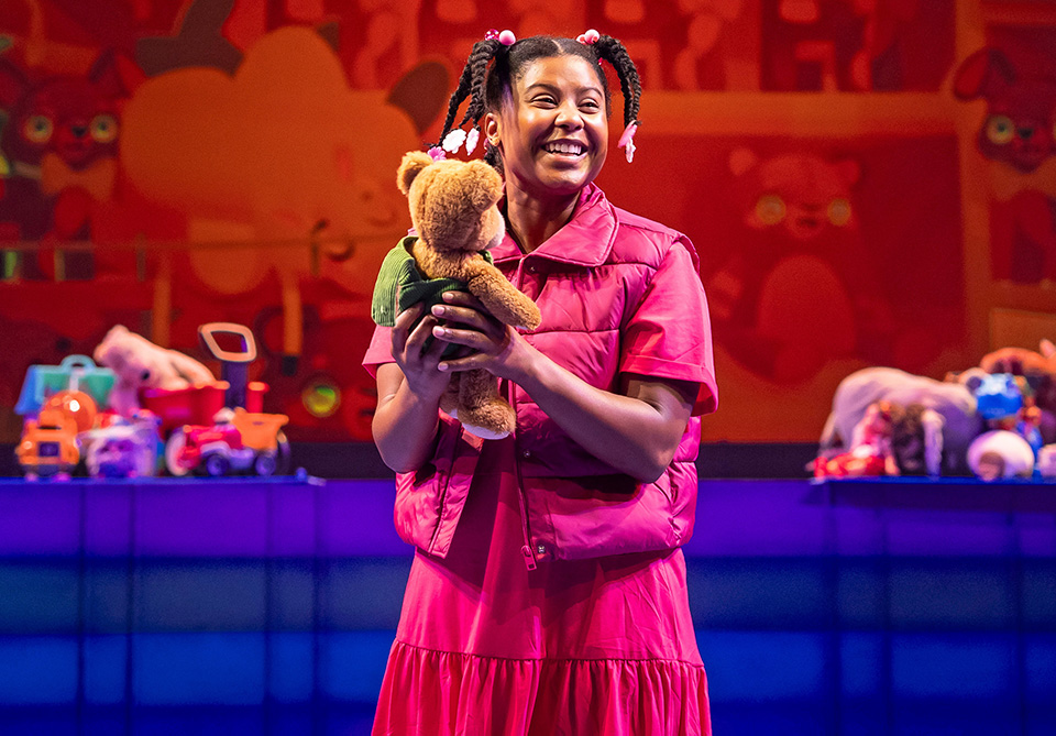 A smiling young girl in a pink outfit holds a teddy bear, surrounded by colorful toys on a brightly lit stage.