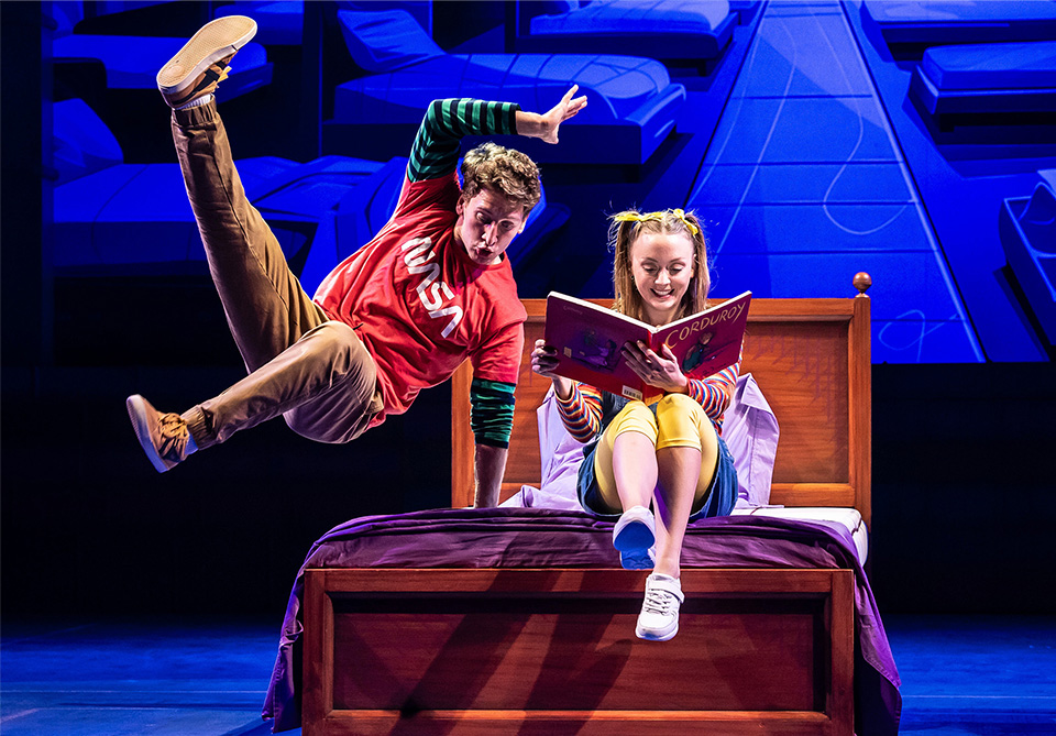 Person in a red NASA shirt leaps in the air next to a young person sitting on a bed reading a book titled "Corduroy."