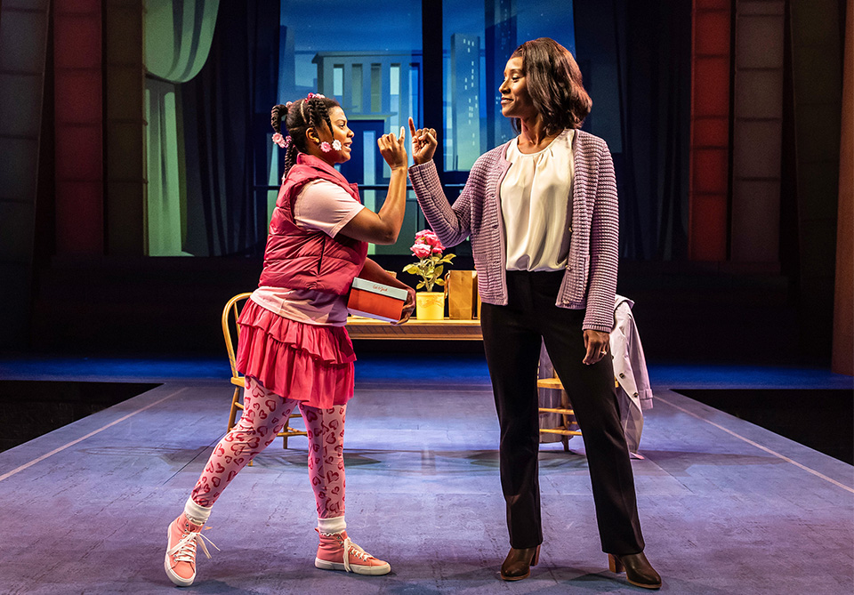 Two actors on stage connect pinky fingers. One is dressed in pink; the other is in a white blouse and lavender cardigan. A table with pink flowers is in the background.