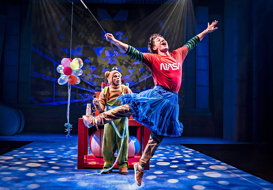 Person in a bear costume holding balloons and another person in a red NASA shirt and blue tutu being lifted in the air.