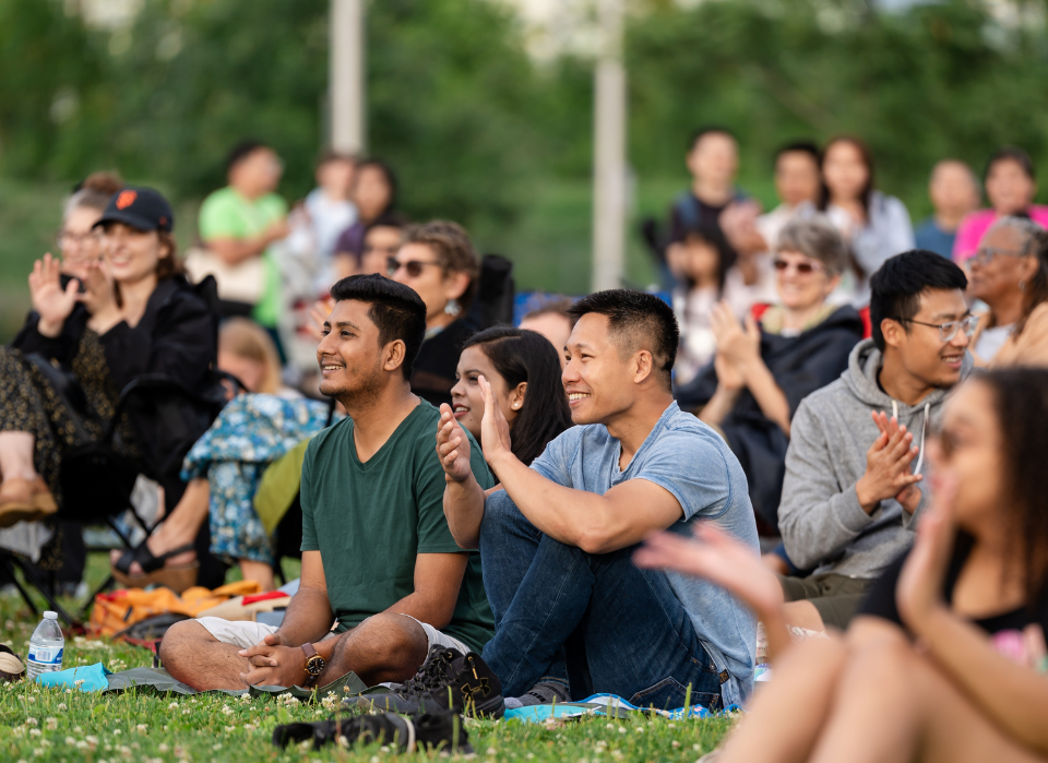 A diverse group of people sitting on grass, smiling and clapping, enjoying an outdoor event. The background is blurred, showing more attendees and trees, indicating a lively and cheerful atmosphere.
