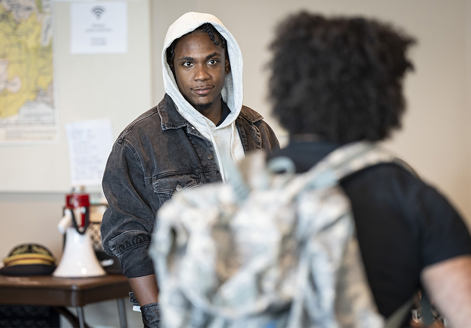 Two people face each other in a room; one has a camouflage backpack and the other wears a denim jacket and white hoodie.