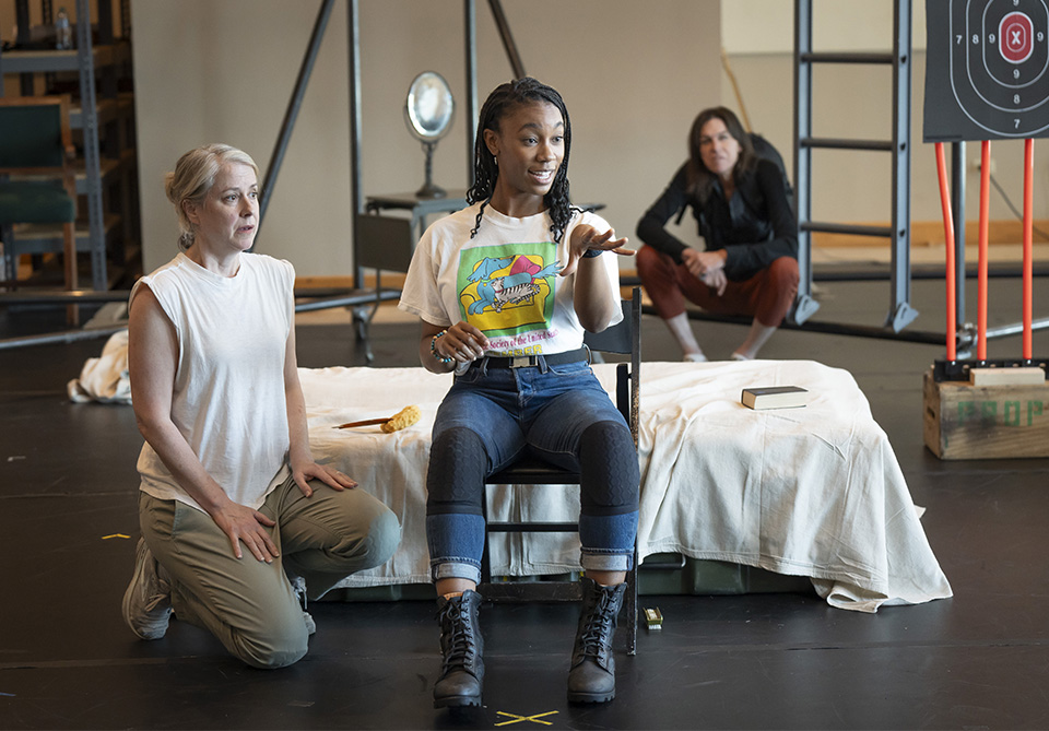Three women are interacting in a rehearsal space with stage props and a target in the background.