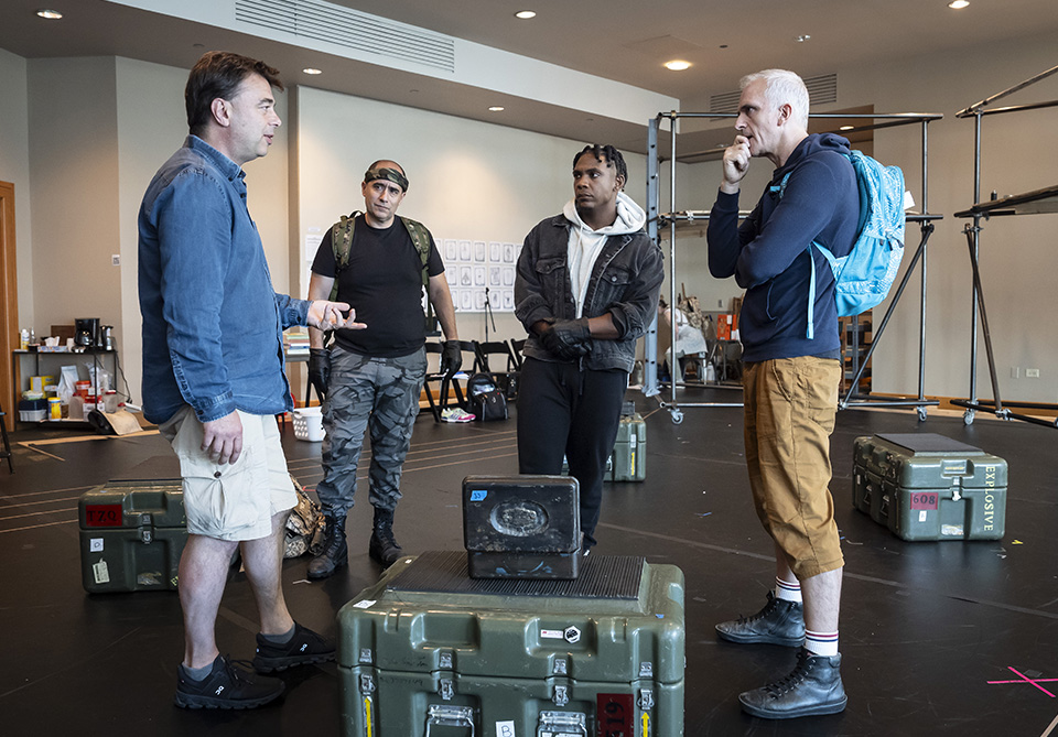 Four men in a rehearsal space, surrounded by military-style crates, appear to be in discussion.