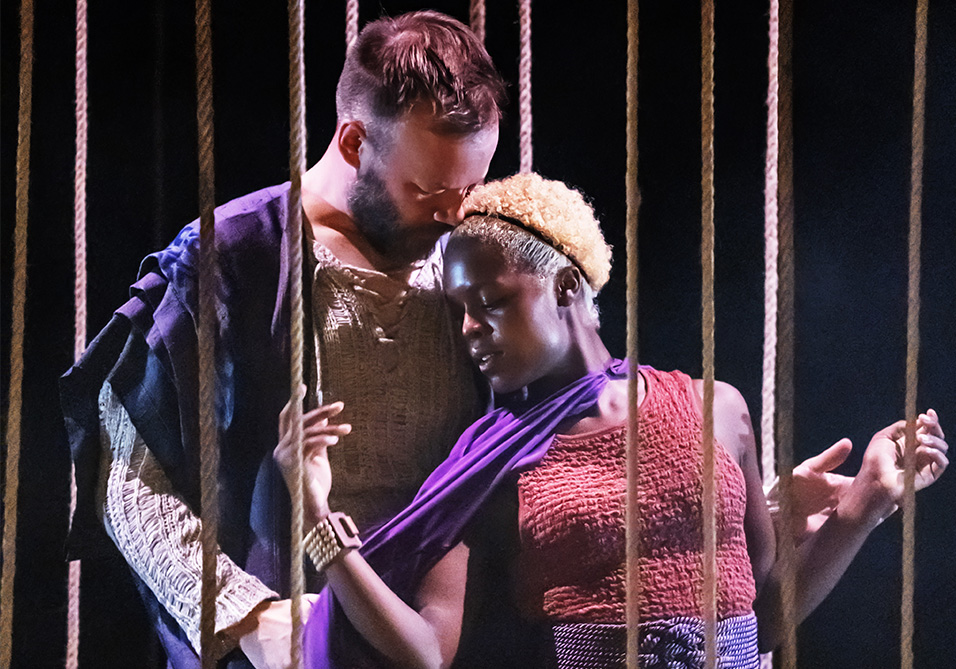 Two individuals in an intimate pose behind vertical ropes, one with short hair and a beard leaning forehead against the other’s head.