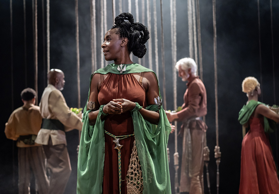 A woman in a reddish-brown dress with a green cape stands on a dimly lit stage, with four other people in the background.