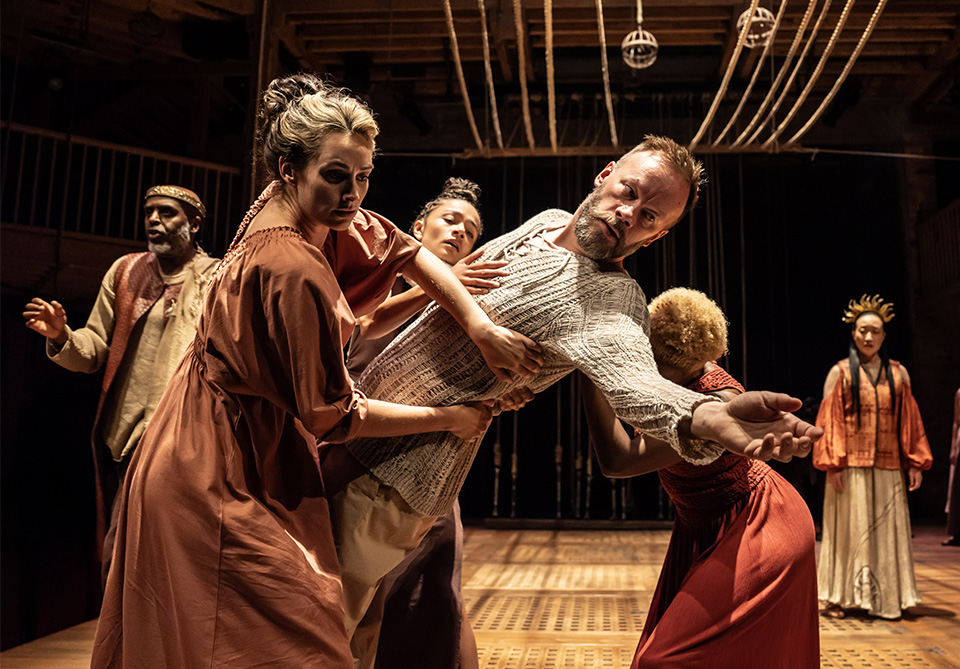 A dramatic scene from a theater performance with a central male actor being supported by three women.