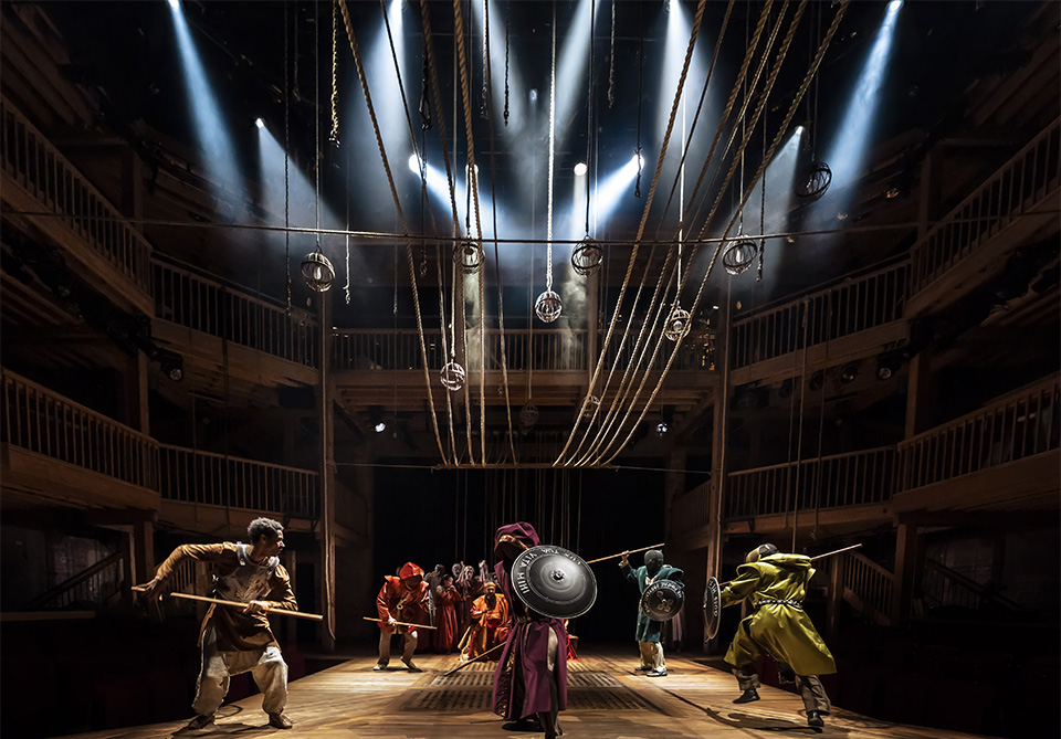 Performers in colorful costumes with props such as shields and staffs on a stage with dramatic lighting and suspended ropes.