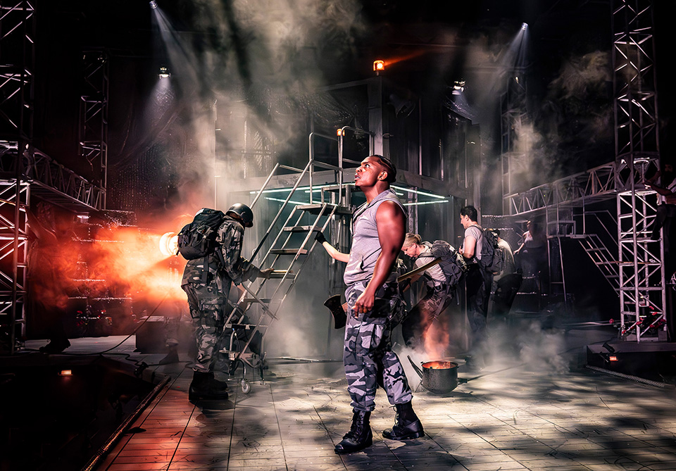 A dramatic stage scene featuring five individuals in military attire with smoke and fiery effects.