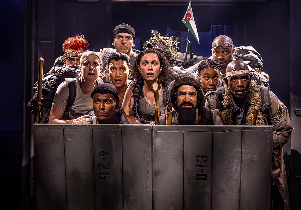 Nine people with intense expressions stand closely together behind metallic crates, two holding wooden sticks, with a dragon-emblazoned flag above.