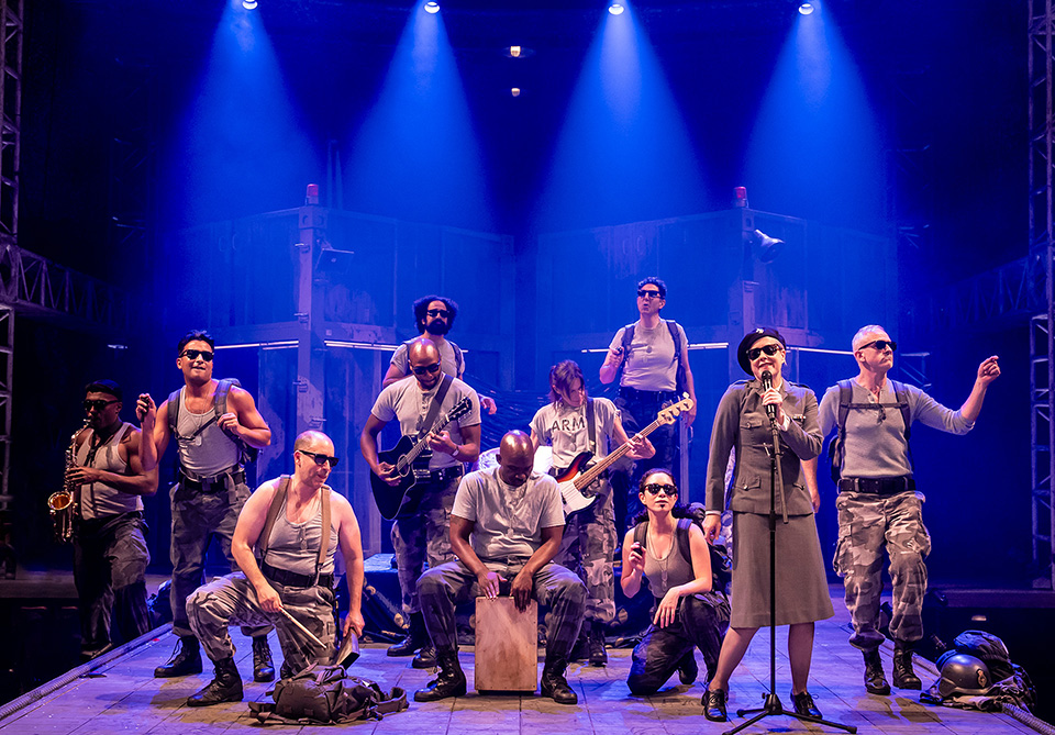 A group of twelve performers in military-inspired attire on stage under blue and purple lights.
