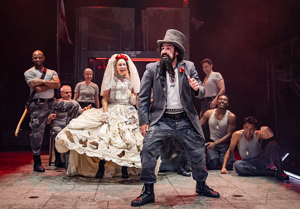 Actors on stage in eclectic costumes, with a man in a top hat and a woman in a tattered wedding dress at the center.