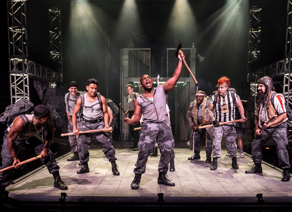 Actors in military-style outfits performing energetically on stage with prop weaponry.