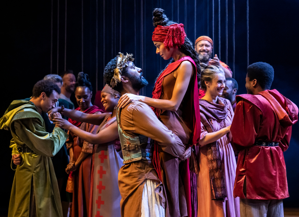 An ensemble of actors dressed in Greek clothing, with man in center holding up a woman in his arms