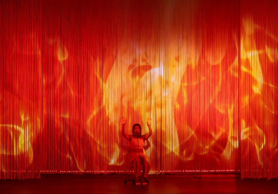 Person seated with raised hands against a curtain backdrop of projected flames in red and orange.