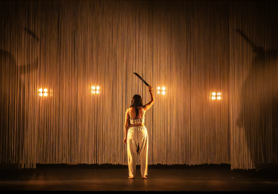 A woman on stage holds a raised machete-like object in front of a lit backdrop of vertical strands and lights.