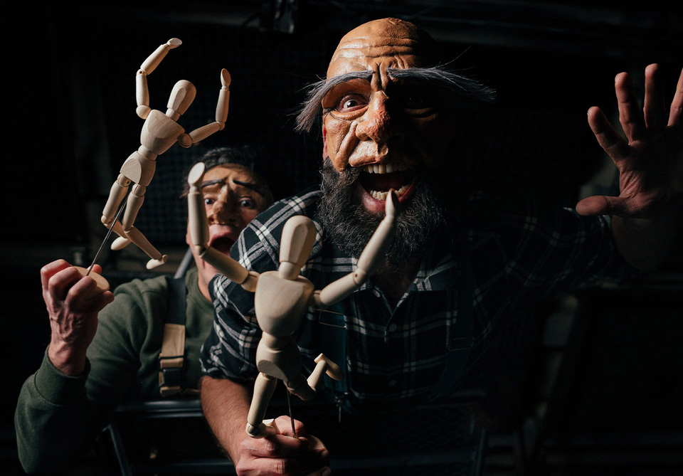 Two people hold and animate wooden artist mannequins; one wears a large mask with exaggerated features.