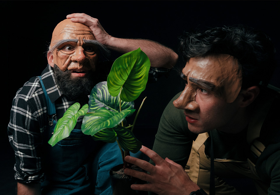 Two people in theatrical masks examine a potted plant with large green leaves.