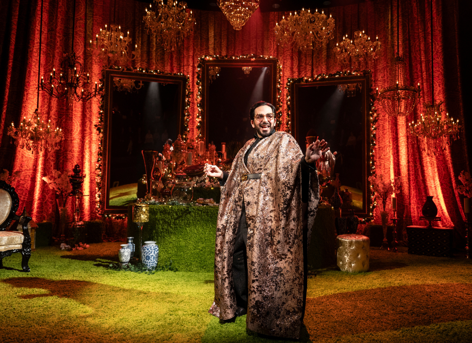 A person in a patterned robe standing in a richly decorated room with red drapes, chandeliers, and mirrors.