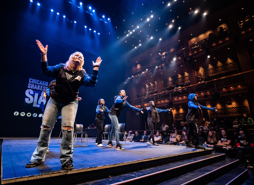 Performers dancing and clapping energetically on a stage with blue and white lighting in a theater.