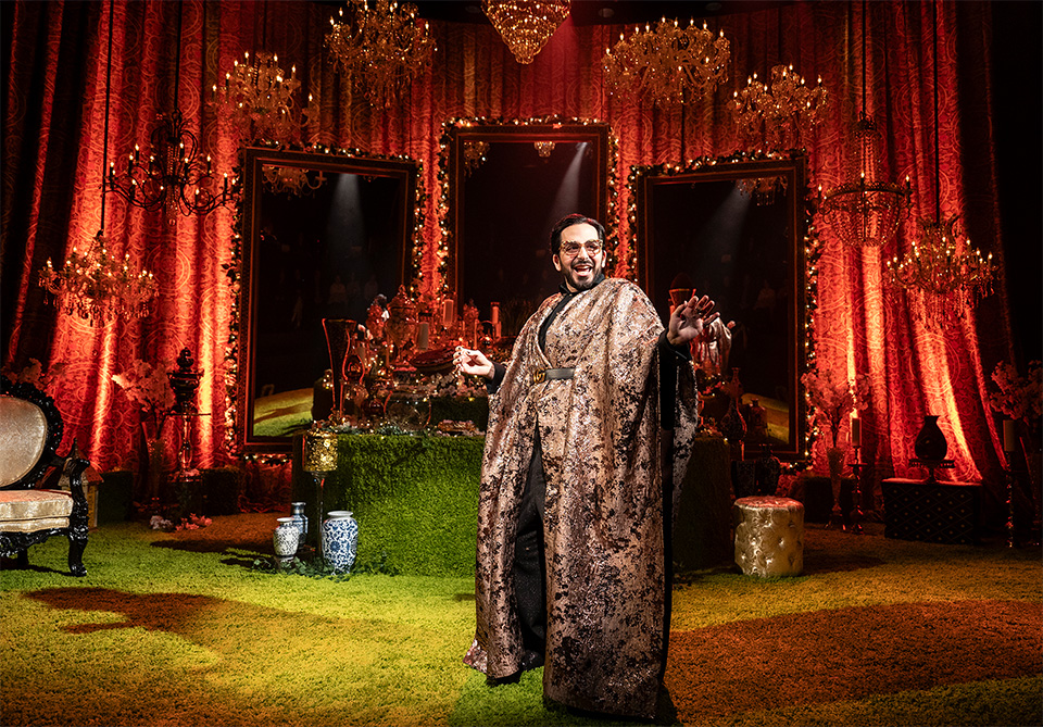A person in an ornate robe stands in a lavish setting with red curtains, mirrors, chandeliers, and decorative objects.