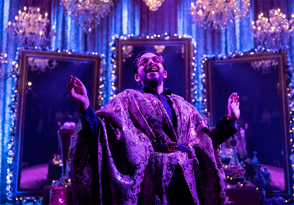 Person in a shimmering cape, surrounded by chandeliers and ornate mirrors, with purple and pink lighting.