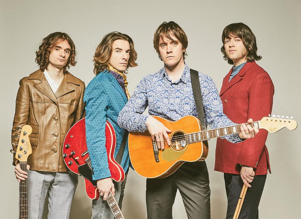 Four men holding musical instruments against a neutral gray background.