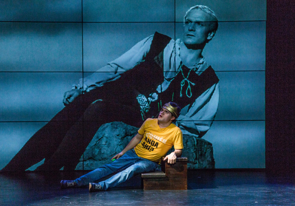 A person in a T-shirt and jeans sits on stage, looking at a black-and-white image of a costumed actor on a large screen behind them.