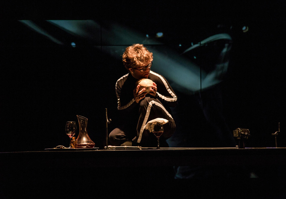 Person in black outfit with white stripes holding a skull on a dimly lit stage, with a wine glass and decanter nearby.