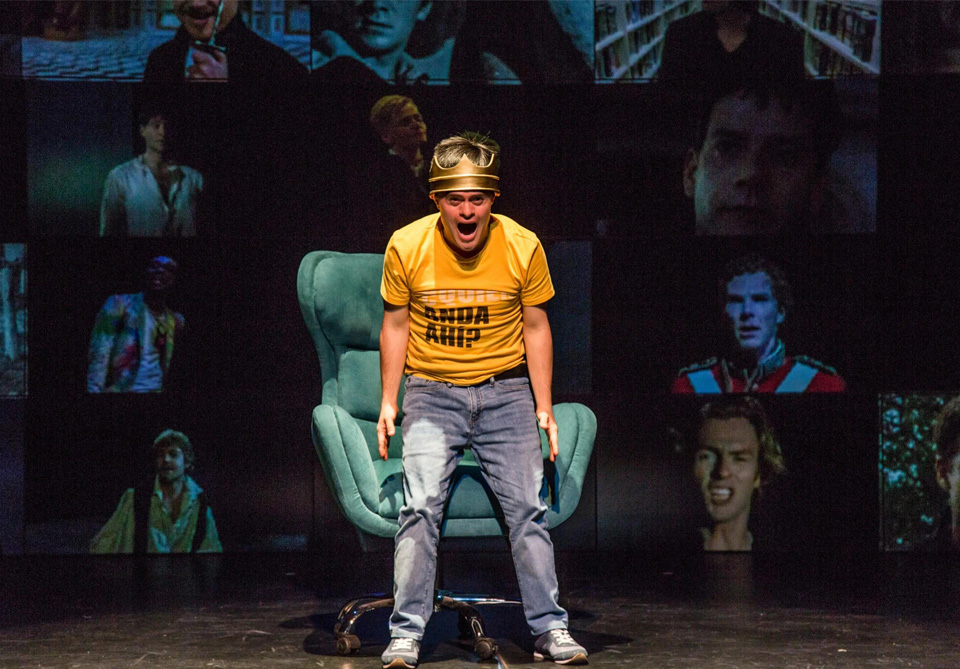 Person in yellow t-shirt and gold helmet stands on stage with a teal chair behind them against a background of screens displaying images.