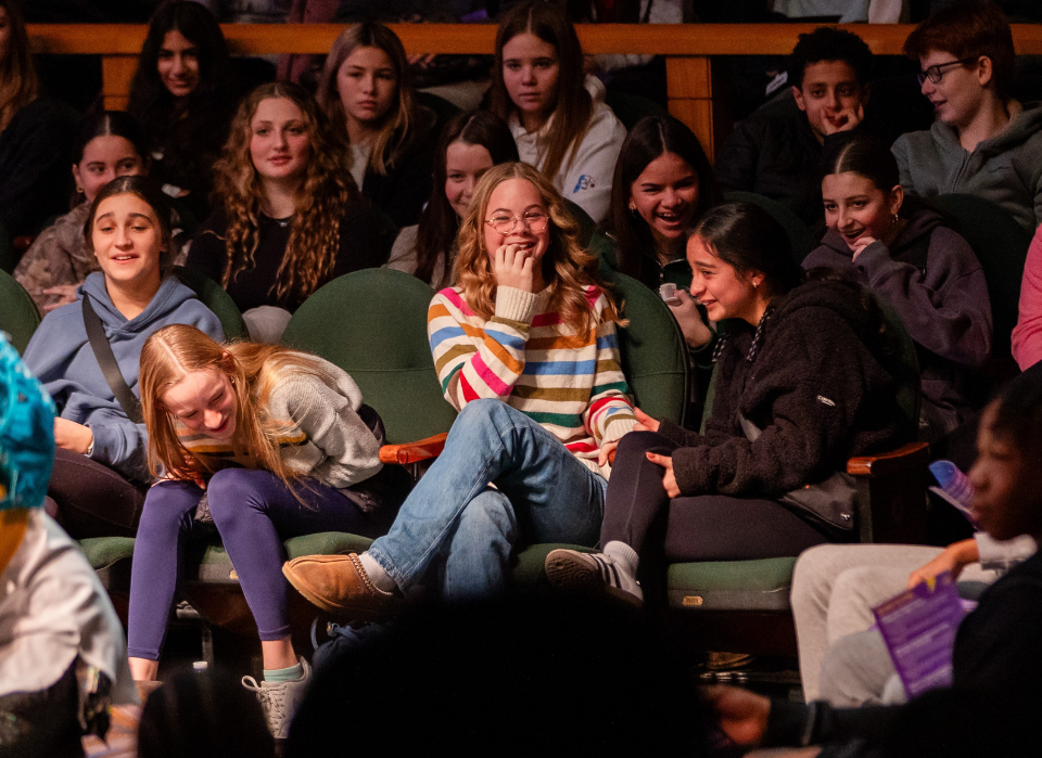 Students in theater seats watching a play, laughing, giggling, and smiling. 